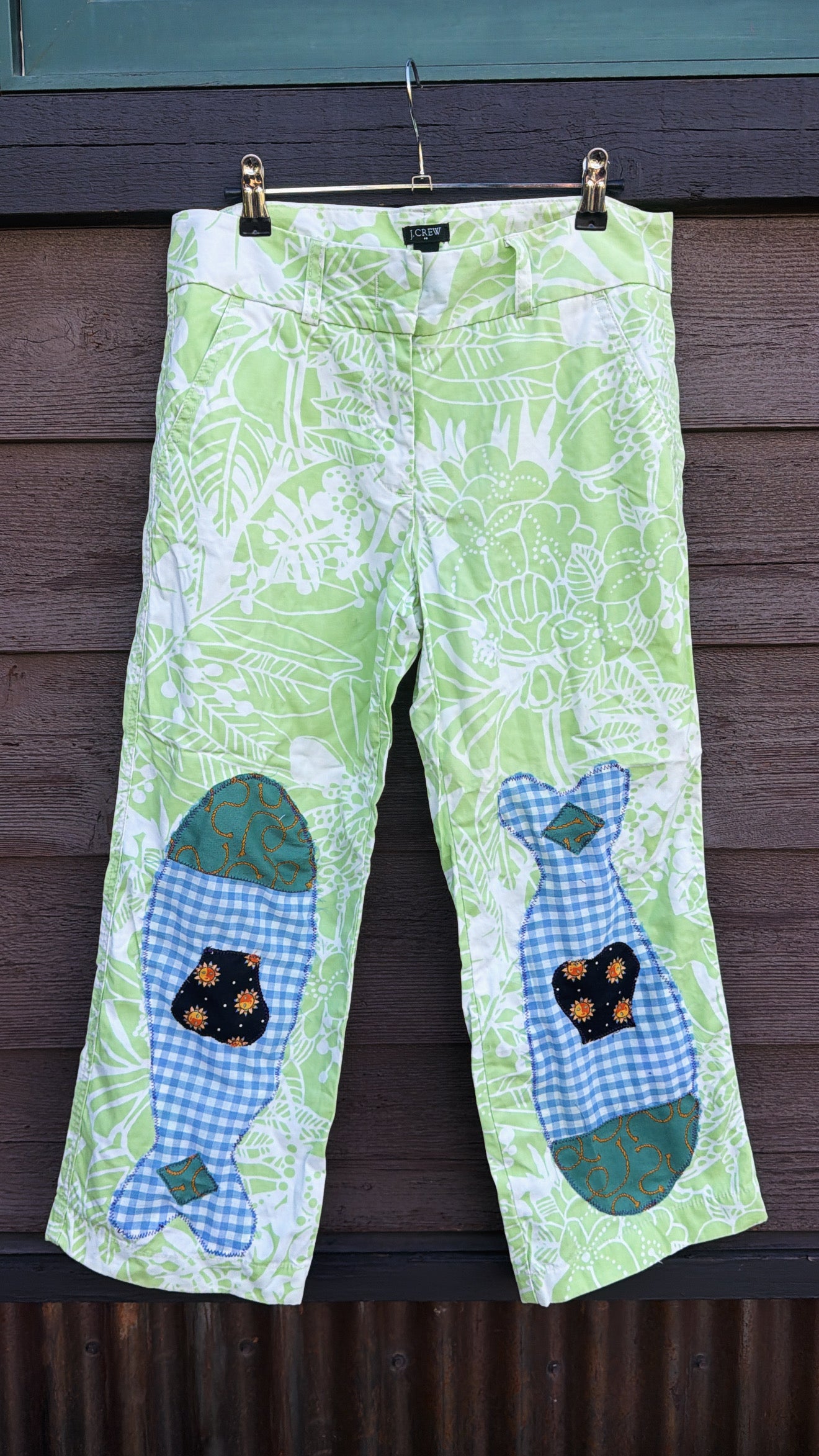 Each fish is pieced together from fabric scraps — blue gingham, teal scroll print, and a navy sun-print cotton — and stitched down with visible decorative stitching that celebrates the handmade process. The matching fish on each leg give them a playful, intentional look that’s anything but ordinary.